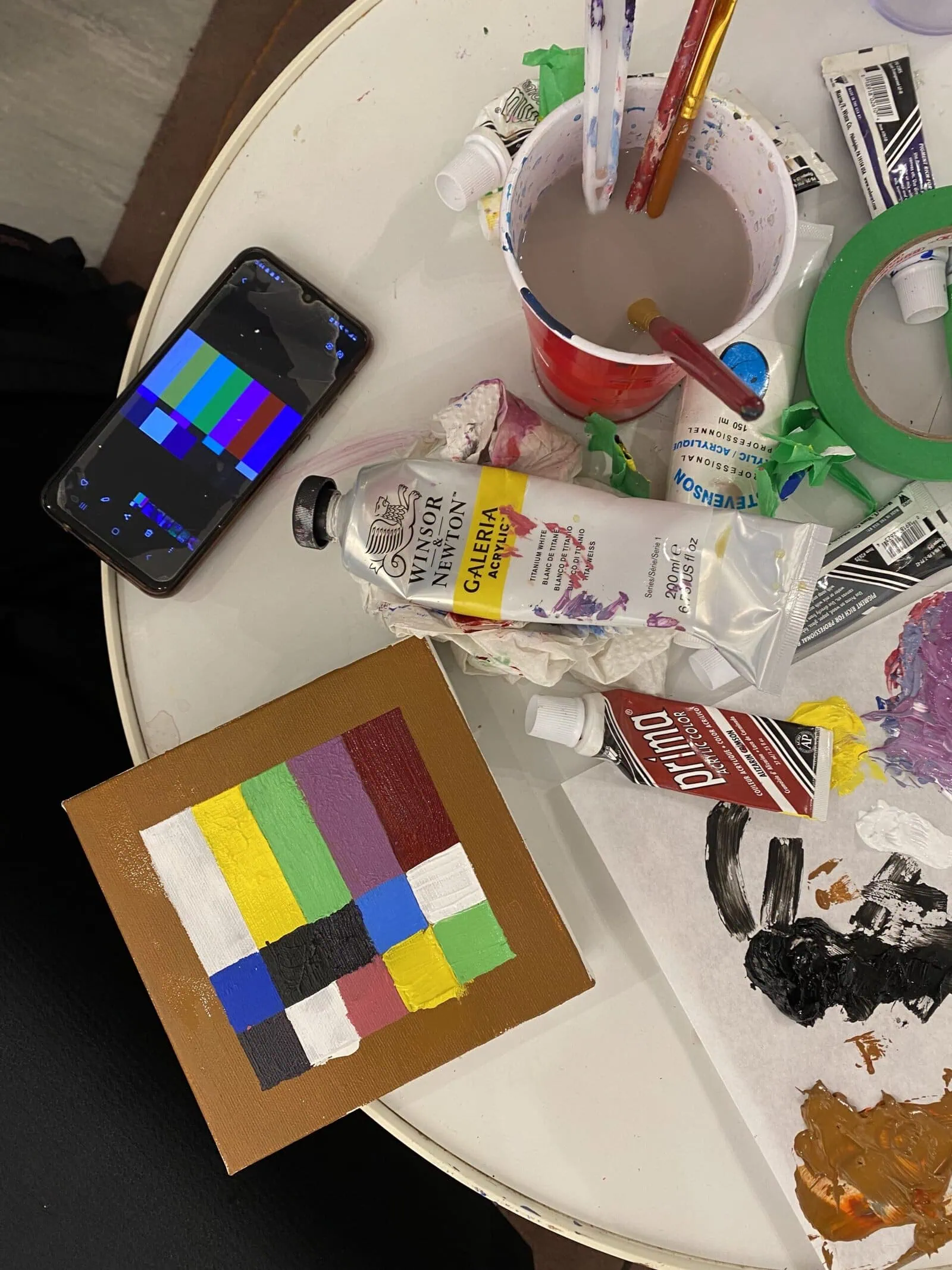 Painting of a television test card with solid-coloured rectangles and squares beside a reference image on a phone and other art supplies.