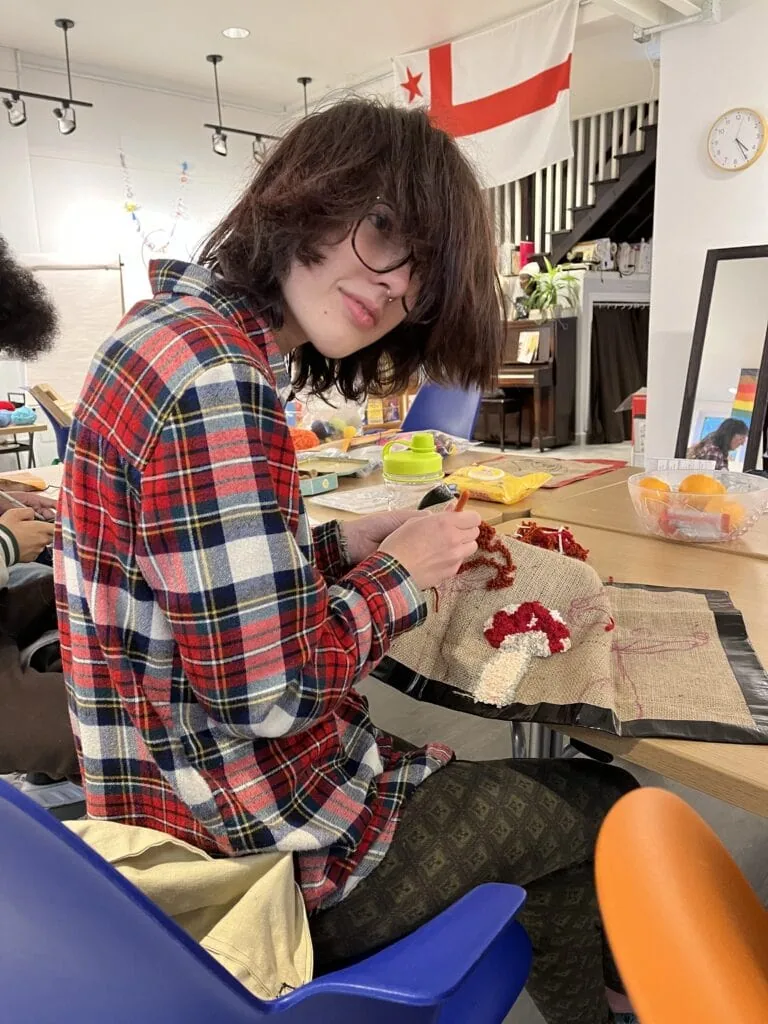 Photo of Caz looking through their long bangs at the camera while rug hooking a mushroom
