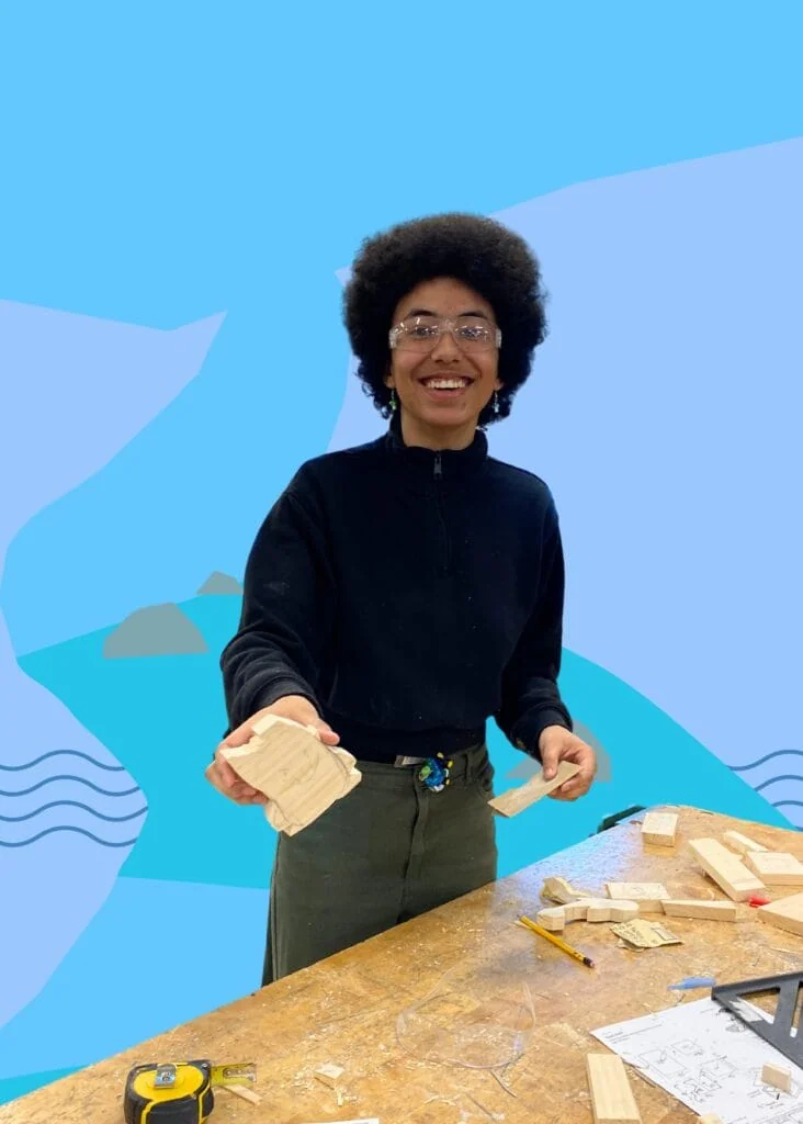 Jayden smiles at the camera while working at a wood working table. The background is blue with waves.