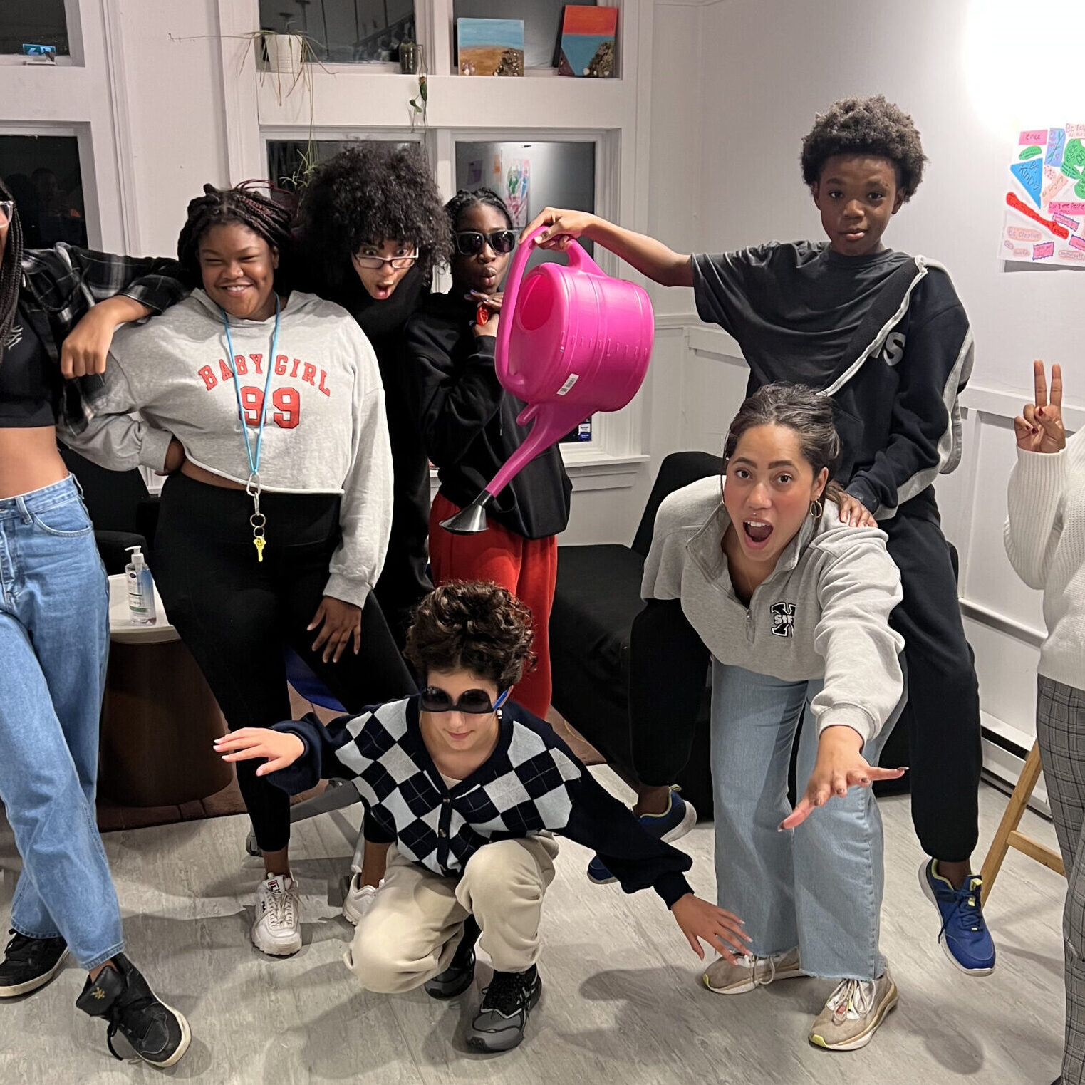 Group of medium and dark-skinned youth strike playful poses indoors with props like a watering can.